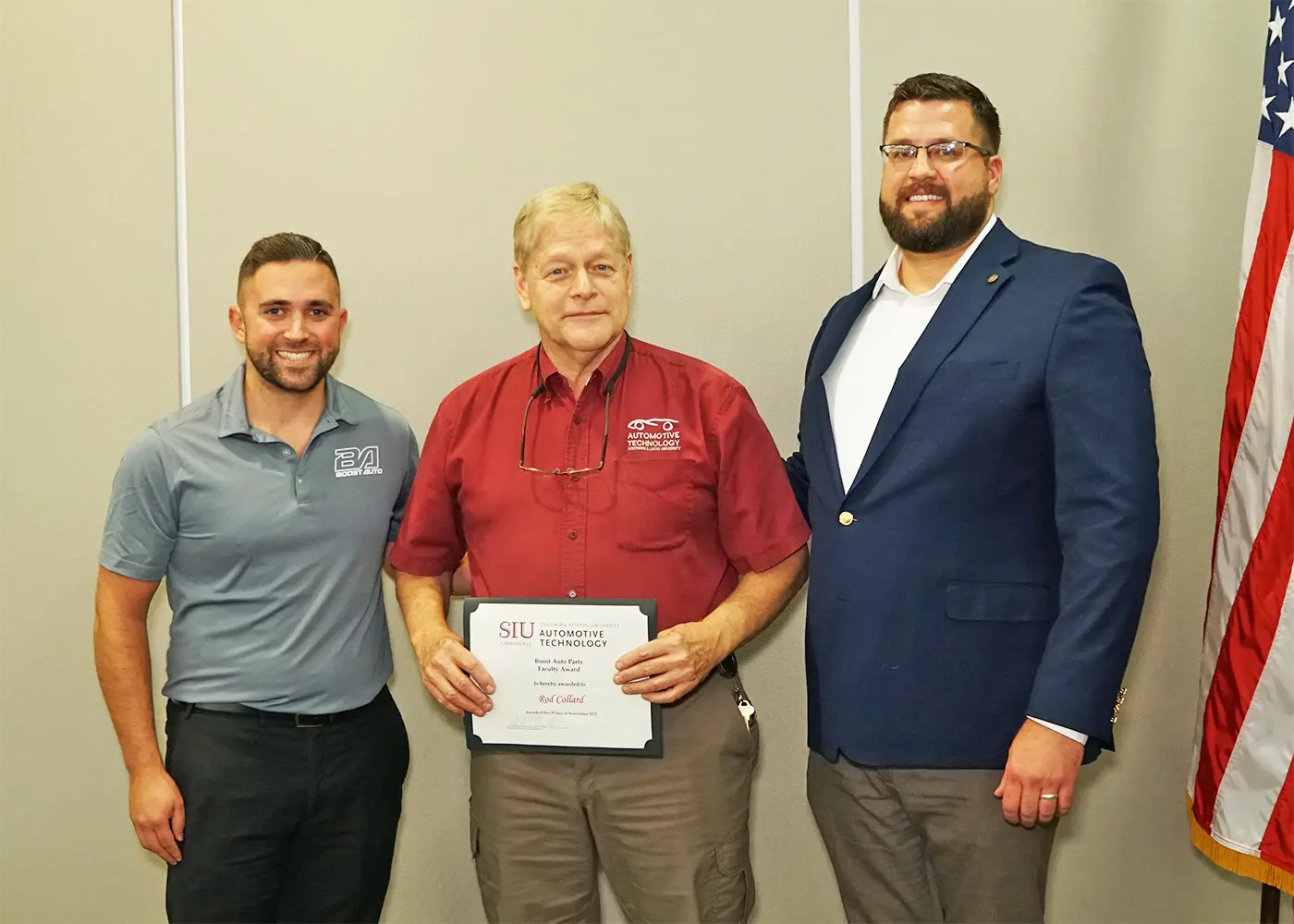 Three men standing together, with one holding an award, in front of a plain wall with an American flag on the side.